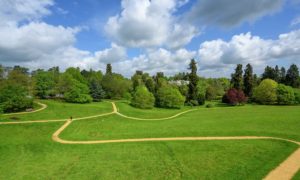 Photograph of a grassland park, criss crossed with surfaced paths. Trees flank the far slopes and large mansion house is just visible.